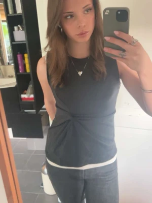 Mörkgrå ärmlös topp med twist - Snygg mörkgrå ärmlös topp från H&M med twistad detalj framtill. Toppen har rund halsringning och är gjord i mjuk bomullsmix. Perfekt att styla med jeans eller kjol för en clean look. Använd fåtal gånger 