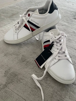 Vita sneakers från Tommy Hilfiger - Snygga vita sneakers från Tommy Hilfiger med klassiska röda, blå och vita ränder på sidan. Skorna har snörning, rund tå och är tillverkade i slätt skinn. Svart detalj på hälen och diskret logga på sidan. Perfekta till jeans eller shorts. Nypris 1200 kr och dessa är sprillans nya.