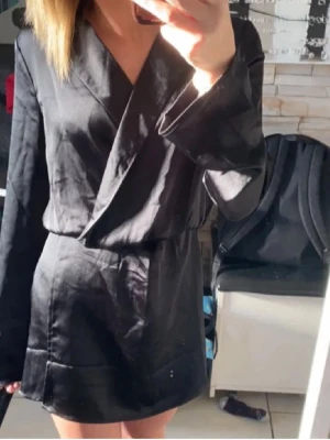 Svart omlottklänning i satin - Säljer en svart kort omlottklänning med långa ärmar i glansigt satinliknande material. Klänningen har en stilren och enkel design med markerad midja och breda slag vid halsen. Perfekt för dig som gillar en elegant och trendig look.