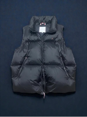 Svart dunväst från Tommy Hilfiger - Snygg svart dunväst från Tommy Hilfiger med hög krage och två dragkedjeförsedda fickor. Västen har TH Protect, vilket innebär att den är vattenavvisande och fylld med dun. Klassisk design med diskret logga på bröstet och dragkedja framtill.