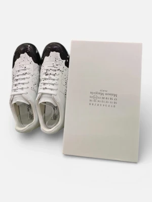 Maison Margiela sneakers med svart stänk - Unika vita sneakers från Maison Margiela i skinn med svart färgstänk och svart detalj på tån. Klassisk låg modell med snörning och rund tå. Snygg kontrast mellan det vita och svarta, perfekt för dig som gillar statement-plagg.