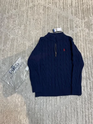 Mörkblå stickad tröja Polo Ralph Lauren - Säljer en mörkblå stickad tröja från Polo Ralph Lauren i storlek M. Tröjan har kabelstickat mönster, half zip-dragkedja och en röd broderad logga på bröstet. Ribbad krage, ärmslut och nederkant ger en snygg och klassisk look. Perfekt för kyliga dagar.