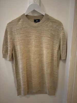 Beige stickad topp från H&M - Säljer en beige stickad topp från H&M med korta ärmar och rund hals. Toppen har en diskret ribbad struktur och är gjord i ett mjukt material som känns skönt mot huden. Perfekt till jeans eller kjol för en enkel och clean look. 
