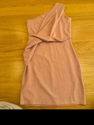 Rosa one shoulder kortklänning - Säljer en snygg rosa kortklänning med one shoulder-design och cool twistad detalj framtill. Klänningen har en tight passform och ärmlös stil, perfekt för dig som gillar trendiga och stilrena plagg. 