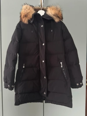 Svart lång pufferjacka med fuskpäls - Säljer en svart lång pufferjacka med stor huva och fuskpälsdetalj. Jackan har dragkedja, tryckknappar, två fickor med dragkedja och justerbara ärmslut. Perfekt för kalla dagar och riktigt snygg till vintern. Jackan ser missfärgad/flammig ut på bilderna men det är tyvärr ljuset som gör det, färgen är jämn och fin i verkligheten. Mer bilder kan skickas vid förfrågan. 