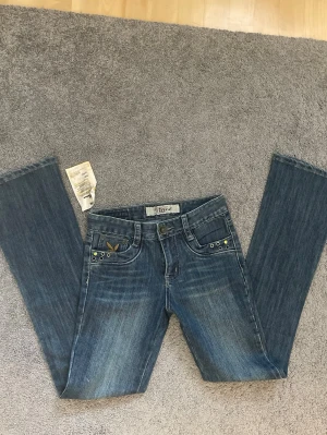 Lowwaist jeans med Playboy-detalj - Säljer ett par blå jeans med bootcut och coola detaljer vid fickorna. På myntfickan finns en broderad gul Playboy-kanin och en guldig sten. Jeansen har låg midja och klassisk femficksdesign. Mått finns på bilderna, skriv vid intresse❤️ Lappar finns kvar 
