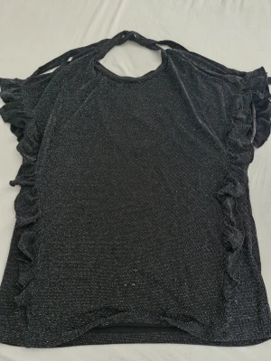 Glittrig svart topp med volangärm från Cream - Säljer en svart glittrig topp med korta volangärmar och knyt i nacken. Tyget har en skimrande effekt och toppen har en rundad halsringning fram och djupare ringning bak. Perfekt för dig som vill sticka ut lite extra. Märke=Cream. Storlek xl, men känns mer som M