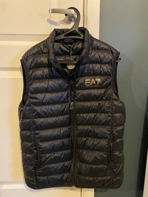 Svart dunväst från Emporio Armani - Säljer en svart dunväst från Emporio Armani EA7 med guldfärgad logga på bröstet. Västen har quiltad design, hög krage och dragkedja framtill. Perfekt att slänga över en hoodie eller tröja för extra värme. Snygg och stilren look. Men de är en XL men den är som en L i storleken. Men jag ska inte betala för frakten.