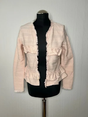 Ljusrosa cardigan jacka med volanger - Säljer en ljusrosa cardigan jacka från Flash Woman i storlek M. Jackan har ett randigt mönster, fransade kanter och volangdetaljer framtill. Materialet är stickat och ger en mjuk känsla. Perfekt för dig som gillar söta och trendiga detaljer.