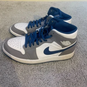 Nike Air Jordan 1 min - Säljer ett par Nike Air Jordan 1 Mid i cementgrått, vitt och blått. Skorna är i riktigt bra skick och är knappt använda. 