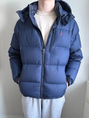 Mörkblå dunjacka från Polo Ralph Lauren - Säljer en mörkblå dunjacka från Polo Ralph Lauren med huva och dragkedja. Jackan har en klassisk puffer-look, diskret logga i rött på bröstet och praktiska fickor med dragkedja. Perfekt för kalla dagar och riktigt skön passform.