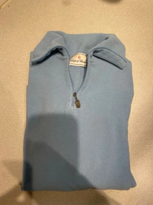 Ljusblå fleece tröja Conte of Florence - Mysig ljusblå fleece tröja från Conte of Florence med halv dragkedja och hög krage. Perfekt för kyliga dagar och enkel att dra på sig. Matchande pannband i samma färg och material ingår.