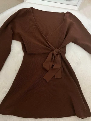 Brun omlottstickad tröja med knyt - Supersnygg brun stickad tröja med omlott och knyt i midjan. Tröjan har lång ärm och v-ringad hals, perfekt för lager på lager under höst och vinter. Materialet är mjukt och stretchigt vilket gör den riktigt bekväm.