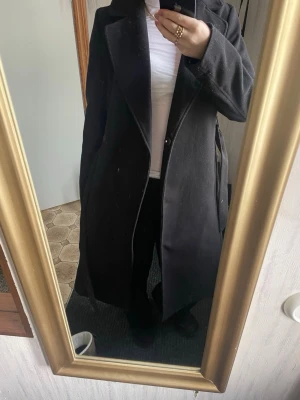 Svart lång kappa med bälte - Säljer en svart, lång kappa med klassisk krage och bälte i midjan. Jackan har en stilren och rak passform, perfekt för dig som gillar minimalistisk stil. Materialet känns tjockt och mjukt, troligen ullblandning. Kappan har långa ärmar och dolda knappar framtill.