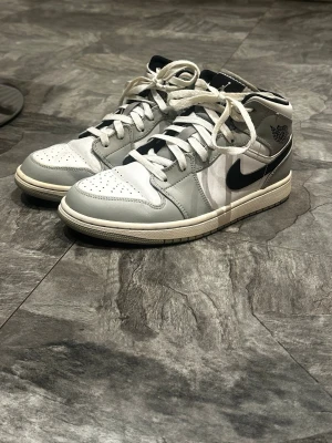 Nike Air Jordan 1 Mid vit/grå storlek 38-39 - Nike Air Jordan 1 Mid i vitt och ljusgrått skinn med svarta detaljer och klassisk swoosh. Skorna har snörning, perforerad tå och ikonisk Air Jordan-logga på sidan. Perfekta för dig som gillar streetstyle och vill ha en clean look.