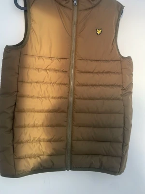 Brun dunväst från Lyle & Scott - Säljer en brun dunväst från Lyle & Scott Junior med gul logga på bröstet. Västen har quiltad design, dragkedja framtill och är tillverkad i syntetmaterial med vattenavvisande yta. Perfekt för lager på lager under höst och vinter.
