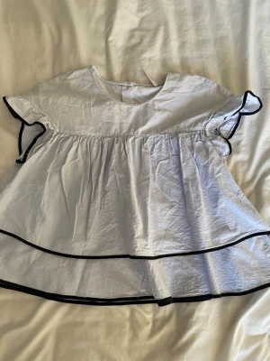 Ljusblå volangblus från Zara - Supersöt ljusblå blus från Zara med volanger och svarta kanter. Blusen har korta ärmar med volangdetalj och knappar i ryggen. Den är luftig och har en lös passform, perfekt för varmare dagar.