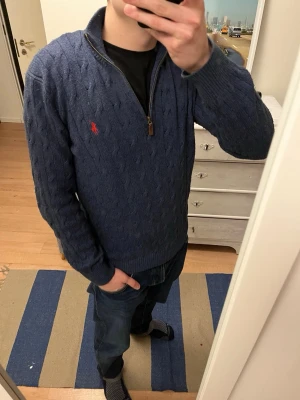 Marinblå stickad tröja Polo Ralph Lauren - Säljer en marinblå stickad tröja från Polo Ralph Lauren med half zip och kabelstickat mönster. Tröjan har långärmad passform, ribbade muddar och röd broderad logga på bröstet. Perfekt för dig som gillar klassisk stil med en modern touch.