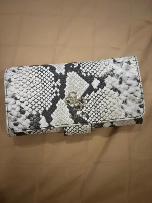 Guess plånbok i ormmönster - Säljer en snygg plånbok från Guess i svartvitt ormmönster med silverfärgade detaljer och logga framtill. Plånboken har flera fack för kort och ID, samt dragkedjefack på baksidan. Materialet är syntet och insidan är silverfärgad.