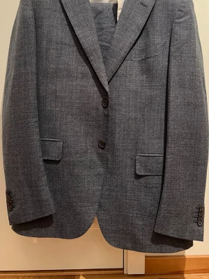 Grå kavaj från Reda Super 110s - Snygg grå kavaj från Reda Super 110s i ull med klassisk passform. Kavajen har två knappar framtill, fyra knappar vid ärmslut och fickor med lock. Fodret är mörkblått och känns lyxigt. Perfekt för dig som vill ha en stilren look. Från Suitsupply, nypris 8549kr