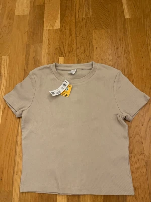 Beige ribbad t-shirt från Lindex  - Säljer en beige ribbad t-shirt från lindex med rund hals och korta ärmar. T-shirten har en clean och enkel look, perfekt för att styla med jeans eller kjol. Materialet är mjukt och stretchigt för skön passform. HELT NY 