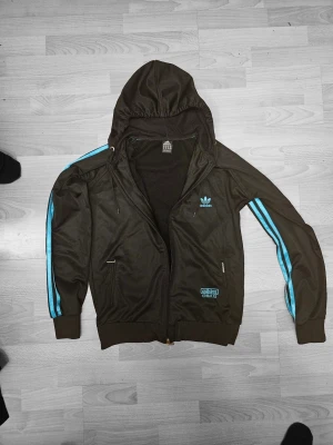 Addidas 2000s vintage zip - Adidas Chile 62 Hooded Track Jacket – Vintage klassiker! Snygg retro Adidas jacka från Chile 62-serien i mörk oliv/svart med starka turkos/blåa detaljer. Heltäckande huva + dragkedja + sidfickor. Klassiska 3-Stripes i neonblått + stort broderat trefoil + "CHILE 62" på bröstet. Riktigt cool 00-tals/vintage känsla – perfekt till streetwear, oversize eller sporty look! På tröjans size står det XL men är liten i storleken och passsar mer som en S