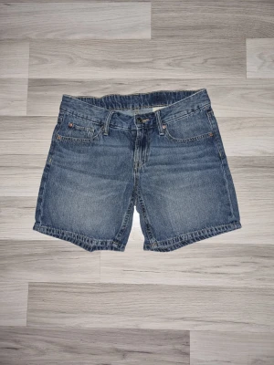 Blå jeansshorts i klassisk modell - Säljer ett par blå jeansshorts från H&M i klassisk femficksmodell. Shortsen har normal passform, bälteshällor och knappgylf. Tillverkade i slitstark bomull med snygga sömmar och raka ben. Perfekta för sommardagar!