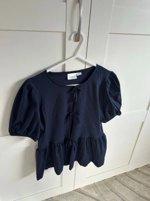 Mörkblå topp med knyt från Gina Tricot - Supersöt mörkblå topp från Gina Tricot med puffiga korta ärmar och tre knyt framtill. Modellen har en lätt volang nedtill och är gjord i mjuk bomull. 