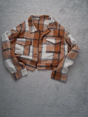 Rutig overshirt från SHEIN XL - Säljer en rutig overshirt från SHEIN i beige, brun och grå. Jackan har stora bröstfickor med lock, knappar framtill och en croppad passform. Perfekt för lager på lager och ger en chill vibe till din outfit.