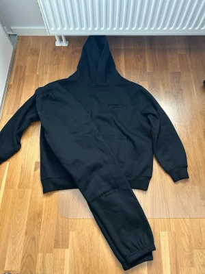 Svart Essentials Fear of God hoodie S - Svart hoodie från Essentials Fear of God i storlek S. Hoodien har stor huva, känguruficka och tryck med logga både fram och bak. Tillverkad i 100% bomull för en skön och avslappnad känsla. Perfekt för dig som gillar streetwear och enkel stil.