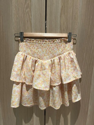 Blommig volangkjol från Kappahl - Supersöt kort kjol från Kappahl med blommigt mönster i ljusa toner av rosa, gul och vit. Kjolen har bred smock i midjan och dubbla volanger som ger en flowy känsla. Perfekt för dig som gillar en lekfull och somrig stil!