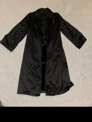 Svart lång morgonrock från Noir - Säljer en svart lång morgonrock från Noir med mjuk, glansig yta och rak passform. Morgonrocken har långa ärmar och är tillverkad i ett material som känns lyxigt och lent. Perfekt för dig som vill ha en stilren och elegant look. Den är silkig i utsidan men har en fluffig insida, perfekt för att hålla en varm.