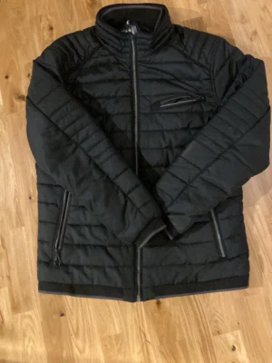 Svart pufferjacka från Calamar - Säljer en svart pufferjacka från Calamar Casual. Jackan har quiltad design, hög krage och tre dragkedjeförsedda fickor framtill. Perfekt för kyliga dagar och har en clean, modern look. Materialet är syntet och jackan är långärmad. Den har 2 innerfickor, en på varje sida (priset går att sänka vid snabbt köp)