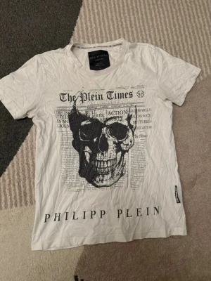 Vit t-shirt med döskalle från Philipp Plein - Säljer en vit t-shirt från Philipp Plein med stor svart döskalle och tidningsinspirerat tryck på framsidan. T-shirten har korta ärmar och rund hals. Märkets logga syns tydligt längst ner på tröjan. Perfekt för dig som gillar edgy streetstyle.