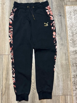 Svarta Puma Kenza mjukisbyxor XS - Svarta mjukisbyxor från Puma x Kenza i storlek XS. Byxorna har blommigt mönster i rosa och rött längs sidorna, guldfärgad Puma-logga på låret och snörning i midjan. Tillverkade i mjukt bomullsmaterial med muddar vid bensluten.