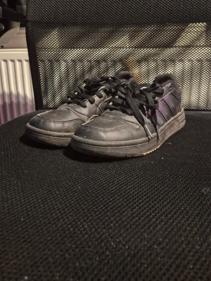 Svarta Adidas sneakers med lila detaljer - Säljer ett par svarta Adidas sneakers med klassiska tre ränder i mörklila på sidan. Skorna har snörning, platt sula och rund tå. Ovandelen är i syntet och textil, vilket gör dem både snygga och bekväma till vardags. Adidas-logga syns på hälen. Har haft de i ett år och de andväns inte längre. Kan tvättas innan de skickas!