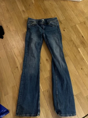 Blå bootcut jeans med fickdetaljer - Säljer ett par blå jeans med bootcut passform och snygga slitningar. Jeansen har unika bakfickor med lock och knapp, samt klassisk femficksdesign. Materialet är jeans i bomull och de har en cool tvättad look.