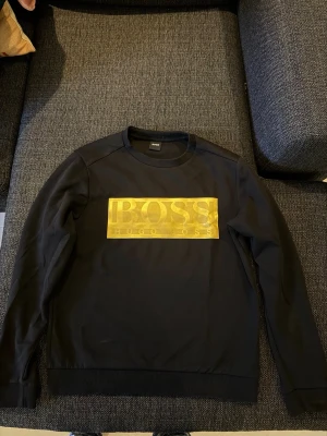 Hugo boss tröja - Jätte fin Hugo boss tröja med stort guld tryck i mitten som gör den unik. Köpt från Zalando. Väldigt fint skicka. Så gott som ny behöver bara stryka den så är den redo. 