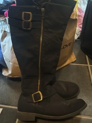 Svarta stövlar med spännen och dragkedja på båda sidor  - Hej Säljer ett par svarta stövlar med dekorativa spännen i mässing och en synlig dragkedja på sidan. Skorna har rund tå, platt sula och är tillverkade i ett material som liknar syntet eller skinn. Perfekta till höst och vinter. Använda en gång har mer ge 