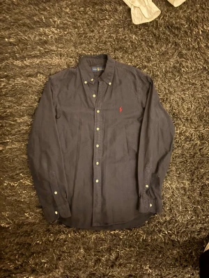 Mörkblå och vit skjorta Ralph Lauren - Två klassiska skjortor från Ralph Lauren, en mörkblå och en vit. Båda har button-down krage, långa ärmar och den ikoniska röda loggan broderad på bröstet. Tillverkade i mjuk bomull och stängs med knappar framtill.