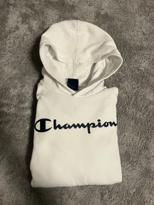 Vit Champion hoodie - Säljer en vit Champion hoodie med mörkblå text. Perfekt till vardag för en chill och skön stil. Storlek 13-14 år mått finns bara att höra av sig! 