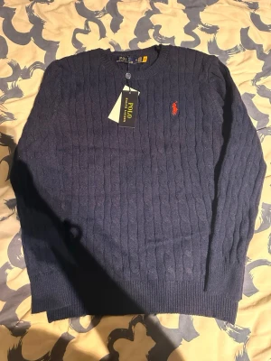 Mörkblå kabelstickad tröja Polo Ralph Lauren - Kabelstickad mörkblå tröja från Polo Ralph Lauren med klassisk röd broderad logga på bröstet. Tröjan har rund halsringning och långa ärmar. Perfekt för dig som vill ha en stilren och tidlös look.