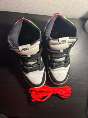 Nike SB Dunk High med färgglatt mönster - Nike SB Dunk High sneakers med svart och vit bas, färgglada grafiska paneler i blått, rött, grönt och gult. Skorna har svart swoosh, svart snörning och extra röda skosnören. Ovandelen är i mocka och textil, med perforerad tå och högt skaft.