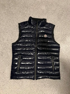 Svart dunväst från Moncler - Säljer en svart, glansig dunväst från Moncler i storlek L. Västen har quiltad design, hög krage, två fickor med dragkedja och klassisk Moncler-logga på bröstet. Perfekt för lager-på-lager och riktigt snygg till streetwear-stilen.