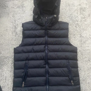 Svart dunväst från BOSS med huva - Säljer en svart dunväst från BOSS med avtagbar huva och två dragkedjefickor framtill. Västen har quiltad design och diskret BOSS-logga på bröstet. Perfekt för lager-på-lager och snygg till streetwear-stilen.