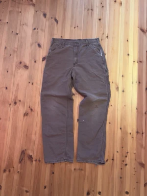 Carhartt Byxor - Säljer ett par bruna byxor från Carhartt med raka ben och flera praktiska fickor. Byxorna har normal passform, bälteshällor och klassisk Carhartt-logga på bakfickan. Tillverkade i slitstark bomull, perfekta för en avslappnad streetwear-look.