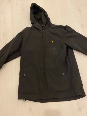Svart vindjacka från Lyle & Scott - Säljer en svart vindjacka från Lyle & Scott med huva och dragkedja framtill. Jackan har två fickor med knappstängning och den klassiska gula loggan på bröstet. Perfekt för blåsiga dagar och enkel att matcha med olika outfits. Den är i nyskick den är bara lite smutsig på bilden 