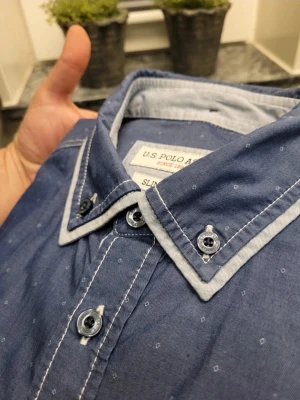 Blå slim fit skjorta från U.S. Polo Assn. - Säljer en blå slim fit skjorta från U.S. Polo Assn. med små diskreta mönster och kontrastdetaljer i vitt på krage och bröstficka. Skjortan har långa ärmar, knappar framtill och broderad logga på bröstet. Perfekt för dig som gillar stilrena detaljer.