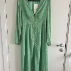 Grön blommig långklänning från Adoore - Superfin långklänning från Adoore i ljusgrönt med små gula blommor. Klänningen har v-ringning, långa ärmar och knappar hela vägen framtill. Tillverkad i ett lätt, glansigt material som känns lyxigt och flowy. Perfekt för vår och sommar!
