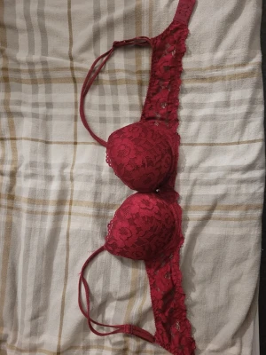Röd push-up bh med rosett och strass - Snygg röd bh med blommig spets, liten rosett och strassdetalj mellan kuporna. Axelbanden är smala och justerbara. Kuporna är vadderade och ger bra stöd. Perfekt för dig som vill ha något extra fint under kläderna. Storlek 75A. 